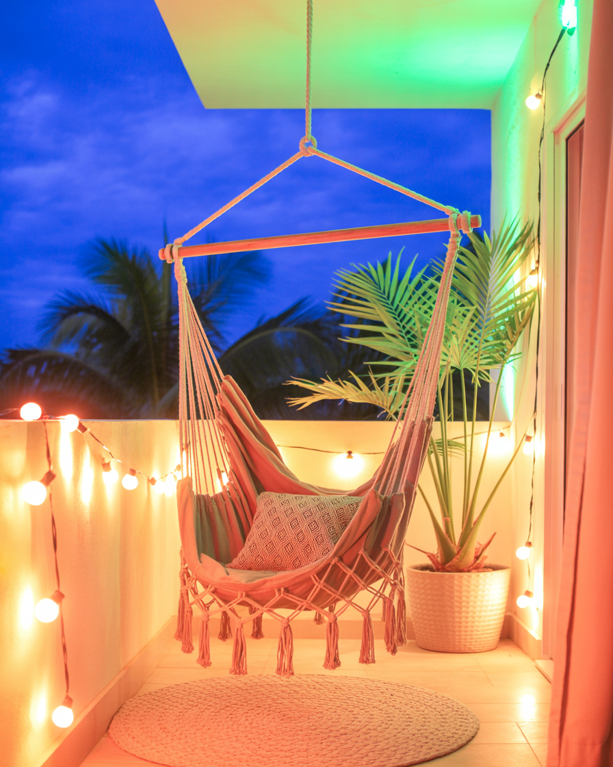 Macramé hammock chair on balcony with string lights and tropical plants at night.