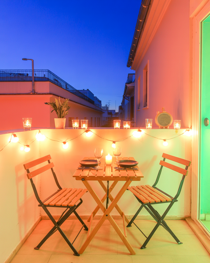 Foldable dining table with dinner setup and string lights on small balcony at twilight.