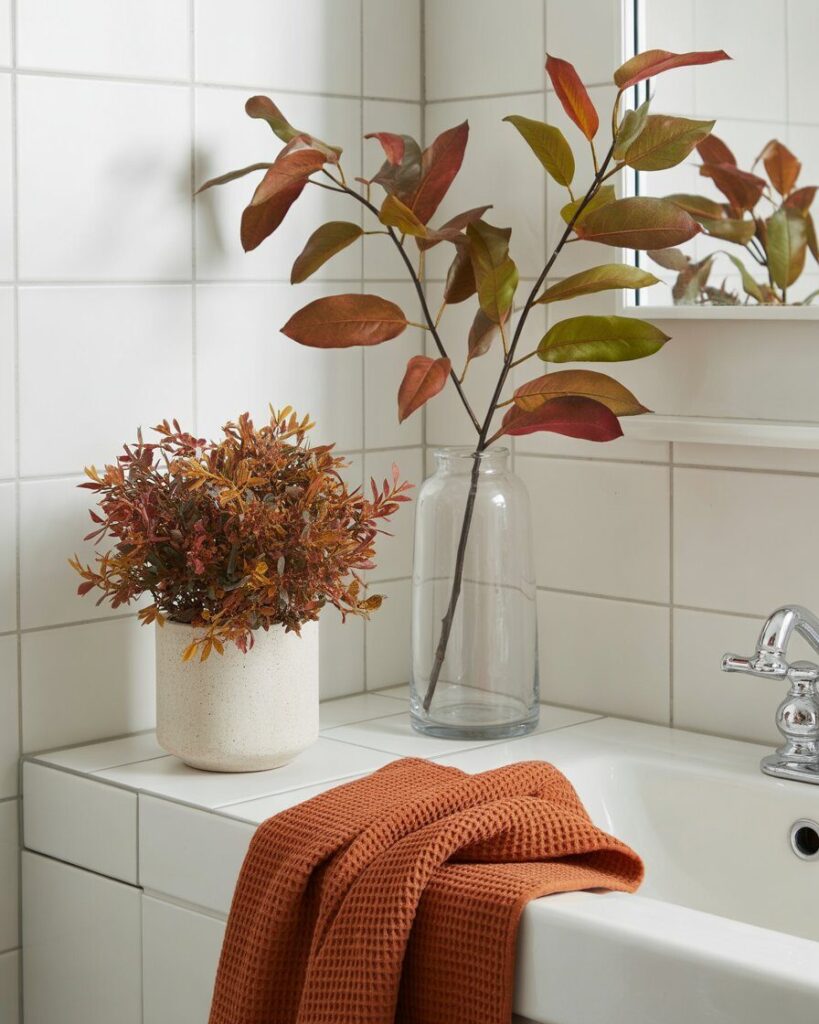 A simple glass vase holding a single, vibrant fall branch with red and orange leaves, placed on a bathroom shelf.